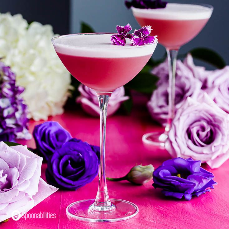Close up photo with an elegant cocktail glass presenting the drink Gin Violette Cocktail garnished with edible flowers. In the background different flowers. Recipe at Spoonabilities.com