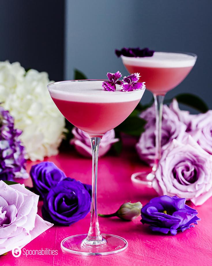 Close up of a cocktail glass with the Gin Violette Cocktail in the center. In the background roses in different purple tones. Recipe at Spoonabilities.com