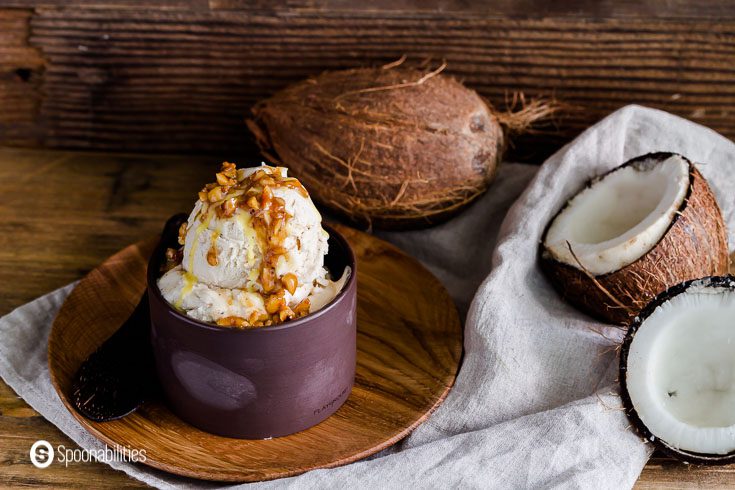 Photo in a rustic/tropical set up with a dark brown bowl on top of a wooden plate with two scoops of ice cream. In the background one whole coconut and another open showing the coconut meat. Recipe at spoonabilities.com