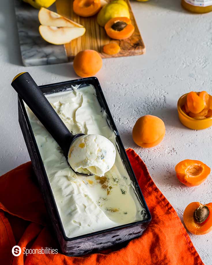 Black ice cream container with a scoop out of Pear Blue Cheese Ice Cream. On the right side some fresh and dried apricots. Recipe at Spoonabilities.com