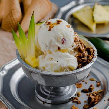 Metal bowl on a metal plate with three scoops of Peppers in Paradise Pineapple Jalapeno Ice Cream. In the background pineapple triangles. Recipe at Spoonabilities.com