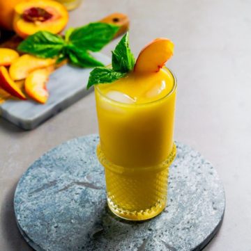A high ball glass with the Tropical Peach Mango Rum Punch, garnished with basil and a sliced peach. The glass in on top of a stone tray. Recipe at Spoonabilities.com