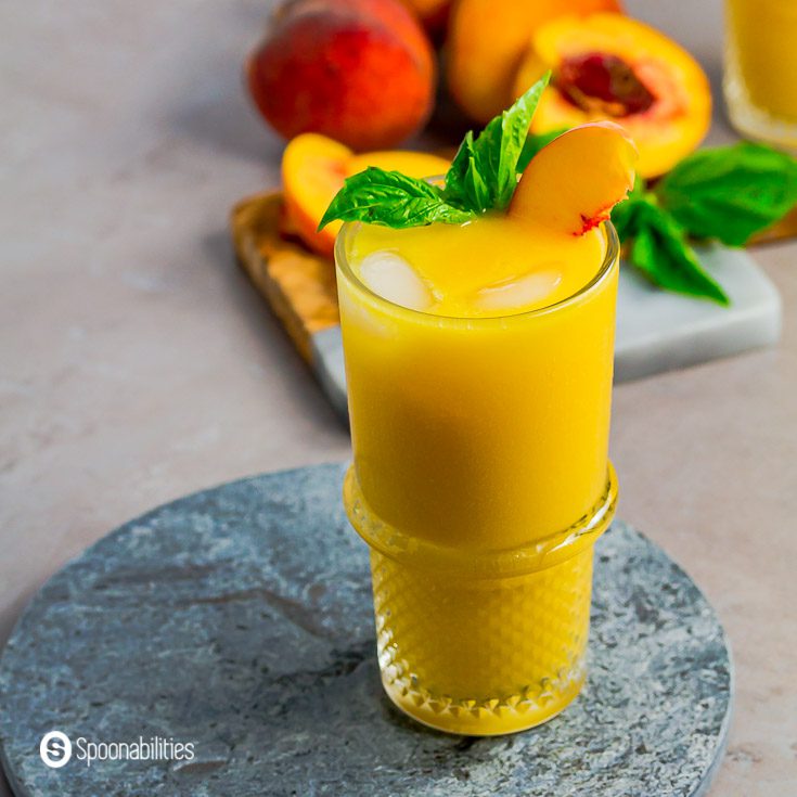 A high ball glass with the Tropical Peach Mango Rum Punch, garnished with basil and a sliced peach. The glass in on top of a stone tray. Recipe at Spoonabilities.com