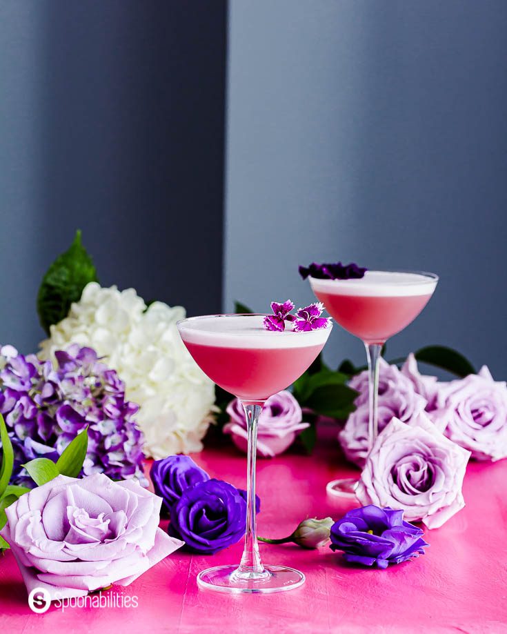 Two cocktail glasses on top of a pink surface with different tones of purple roses around the scene. The two glasses have the Gin Violette Cocktail with a beautiful froth and garnished with edible flowers. Recipe at Spoonabilities.com