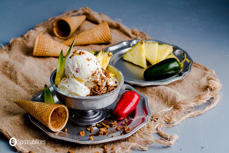 Tropical set up with a burlap napkin. The metal plate in the center has a pewter bowl with three pineapple jalapeno ice cream bowls. In the background a round plate with pineapple triangles. Spoonabilities.com