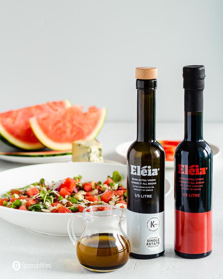 In the background a summer salad and in the front two black bottle and next to them a small glass container with a balsamic vinaigrette. The bottles are Eleia extra virgin olive oil and balsamic vinegar. Products available at Spoonabilities.com