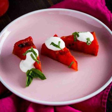 a photo with a pink round plate with three pieces of grilled watermelon topped with Greek yogurt lime crema and garnished with fresh mint. Recipe at Spoonabilities.com