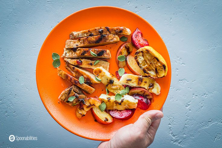 A hand holding a orange round plate with grilled pork chops, plums, halloumi cheese, and grilled lemons. Garnished with fresh oregano. Recipe at Spoonabilities.com