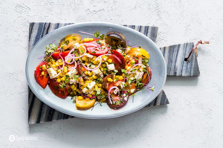 Oval serving plate on top of a marble board with a summer salad with Heirloom Tomatoes, red onion, microgreens, feta cheese, and corn. Recipe at Spoonabilities.com