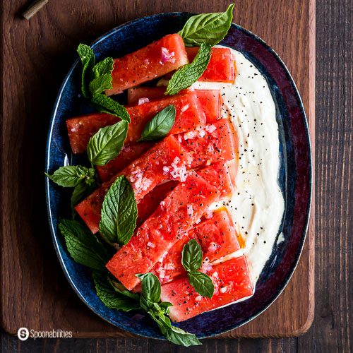 Overhead photo with An oval blue plate on top of a dark wooden board with ten pieces of Marinated Watermelon Salad on a bed of Whipped Feta Dip and garnished with fresh herbs, and Middle-East spice mix. Recipe at Spoonabilities.com