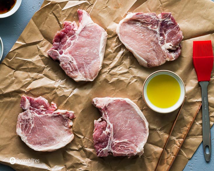 Four raw pork chops on top of a paper bag. Next there is a small pinch bowl with Greek olive oil with a brush. Recipe at Spoonabilities.com