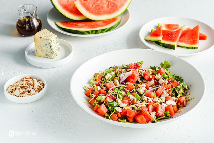 Summer salad in a white serving plate. The salad has watermelon, arugula, blue cheese, almonds, red onions, and dressed with a balsamic vinaigrette. In the background of the photo a small plates with the different ingredients. Recipe at Spoonabilities.com