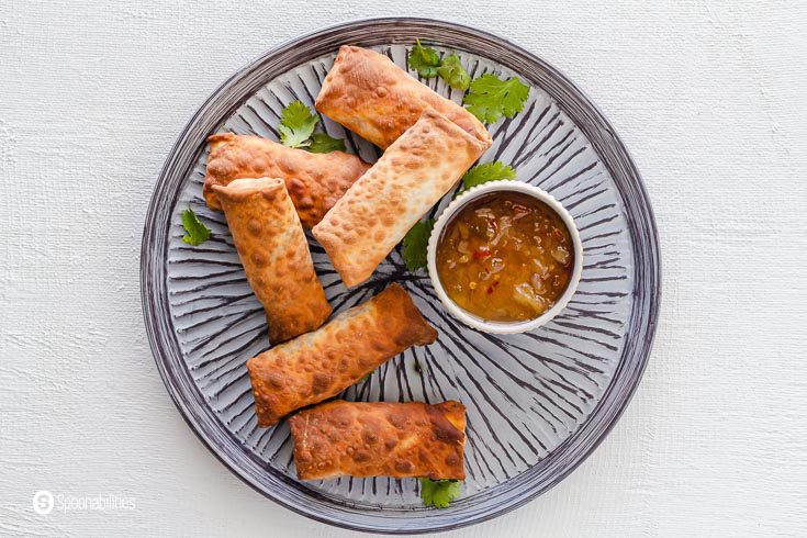 A round grey plate with six Vegetable Egg Rolls and in a small bowl pineapple habanero sauce. Recipe at Spoonabilities.com
