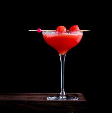 Photo with a black background and a tall glass sitting on a wooden platform. The cocktail glass has a bright red watermelon cocktail and garnished with two watermelon balls. Recipe at Spoonabilities.com