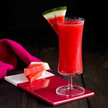 A tall glass on top of a small red porcelain board with two pieces of watermelon. The glass has a Watermelon Mai Tai Cocktail. Recipe at Spoonabilities.com