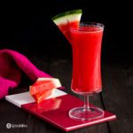 A tall glass on top of a small red porcelain board with two pieces of watermelon. The glass has a Watermelon Mai Tai Cocktail. Recipe at Spoonabilities.com