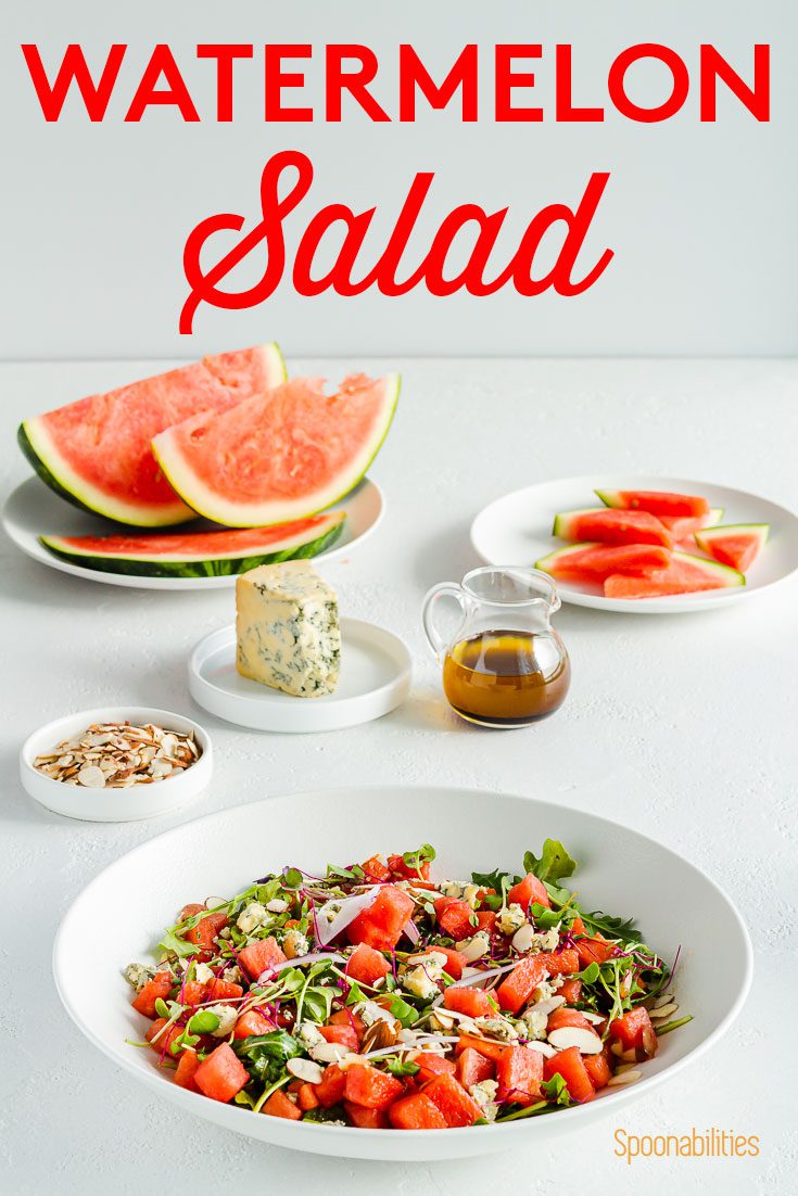 45 degree angle of watermelon salad with blue cheese, red onions, arugula, and almonds. Presented in a Large white bowl in the center and in the background the balsamic vinaigrette, fruit chunks. Recipe at Spoonabilities.com