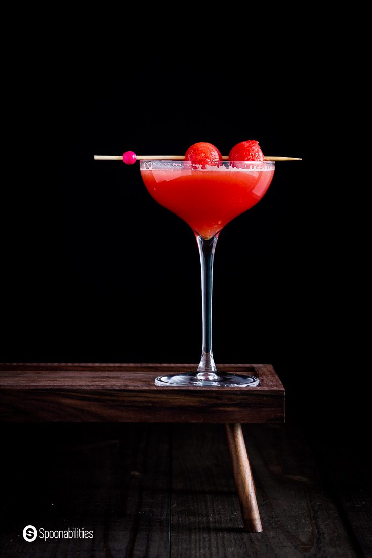 A beautiful tall cocktail glass in a wooden pedestal with a red watermelon cosmo in a dark background. Recipe at Spoonabilities.com