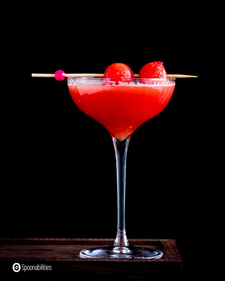 A Close up of this summery and refreshing drink called Watermelon Cosmopolitan. The photo has one tall glass with a drink in a dark background. Recipe at Spoonabilities.com