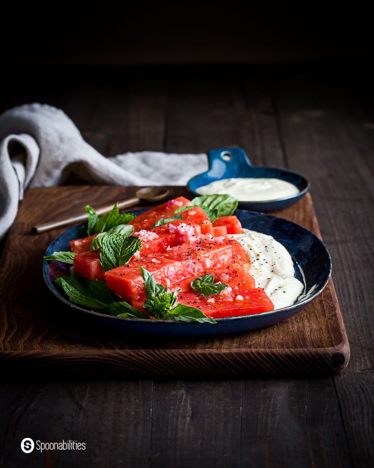 Wooden board with an oval blue plate with marinated watermelon pieces on top of a feta dip. Recipe at Spoonabilities.com