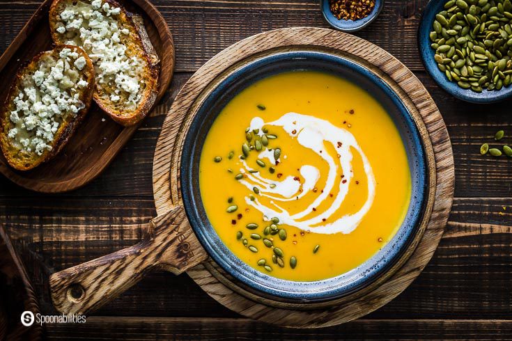 Overhead photo with a blue bowl with butternut squash soup garnished with roasted pumpkin seeds, coconut cream drizzle, and smoked chili flakes. You will see a wooden board with two honey feta toasts around the bowl and on the left side, a pinch bowl with pumpkin seeds. Recipe at Spoonabilities.com