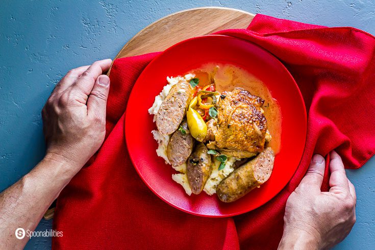 Two hands holding a red plate which is on top of a red napkin. The plate has chicken scarpariello with mashed potatoes. Recipe at Spoonabilities.com