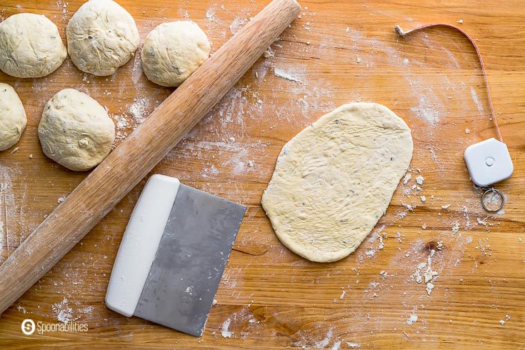 Wooden board with some dough balls and one of the pieces getting the traditional naan shape with a rolling pin. recipe at Spoonabilities.com