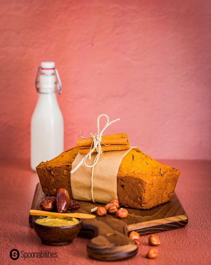 A loaf of bread on top of a wooden board decorated with hazelnuts, dates, and a small dish with brown butter. In the background a bottle with milk. Recipe at Spoonabilities.com