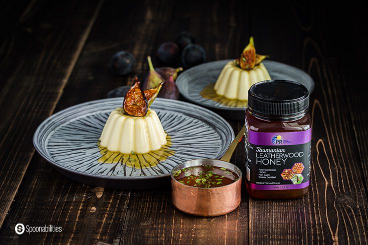 two plates in the background with a Goat Milk Corn Panna Cotta with with Caramelized Figs & Honey Lemon Thyme Drizzle. In the front of the photo is a jar of Tasmanian Leatherwood Honey with a tiny pot with the honey drizzle. Recipe at Spoonabilities.com