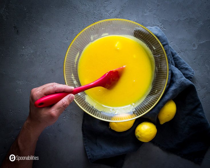 Making the lemon curd: A hand with a red spatula stirring the egg custard with the butter. Steps how to make this lemon curd at Spoonabilities.com