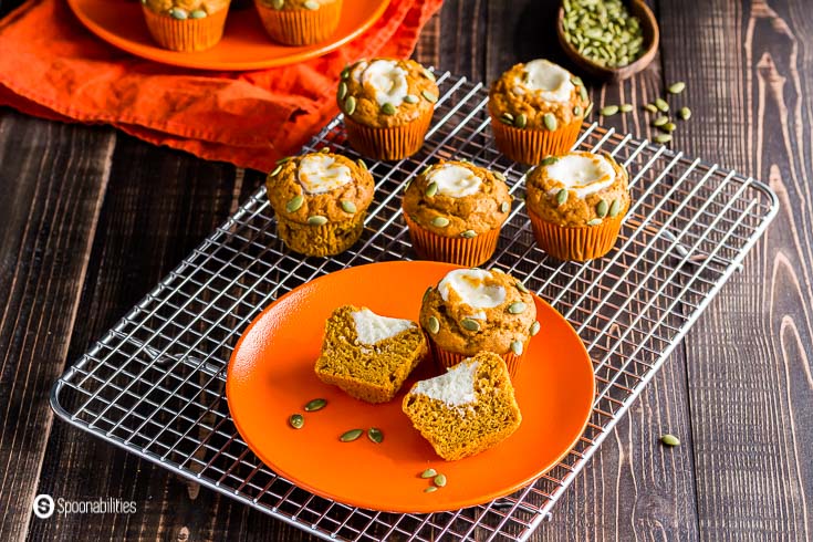 Horizontal photo with several muffins. In an orange round plate two pumpkin muffins and one cut in a half showing the cream cheese filling. Recipe at Spoonabilities.com