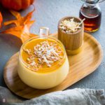 A glass cup with Pumpkin coconut panna cotta. This fall dessert is garnished with a drizzle of cinnamon-infused maple syrup and toasted coconut flakes. Recipe at Spoonabilities.com