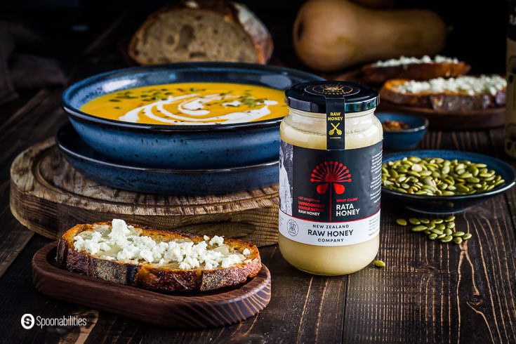 This photo has a jar of the Raw Rata Honey from New Zealand. On the left side, a Feta Honey toast on a small wooden board, and in the background, a soup bowl with butternut squash soup. Recipe at Spoonabilities.com