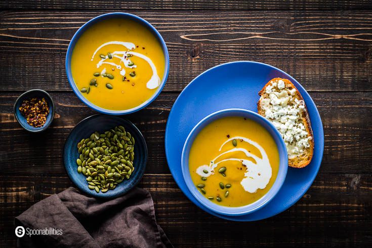 Overhead photo with two soup bowls with butternut squash soup and in other two pinch bowls pumpkin seeds and smoked chili flakes. Recipe at Spoonabilities.com