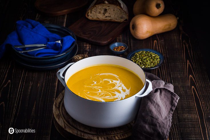 Large white pot with butternut squash soup. In the background, whole butternut squash as décor with some plates and other items. Recipe at Spoonabilities.com