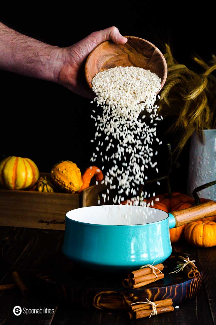 Pouring Arborio rice in a teal color pot with a wooden handler. The table is decorated with pumpkins and fall leaves. Recipe at Spoonabilities.com