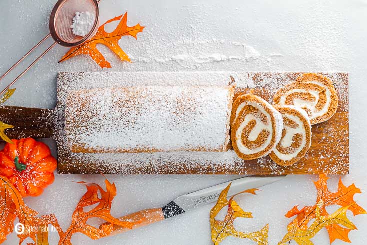 Overhead photo with the pumpkin cake roll on a wooden board, dusted with powdered sugar and decorated with fall leaves and pumpkins. Recipe at Spoonabilities.com