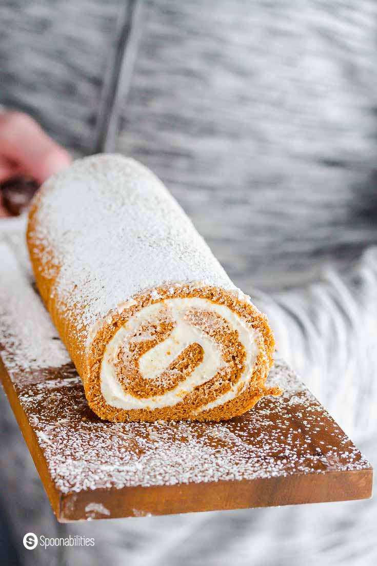 A person with a grey jacket holding the wooden board with the pumpkin cake roll and dusted with powder sugar. Recipe at Spoonabilities.com