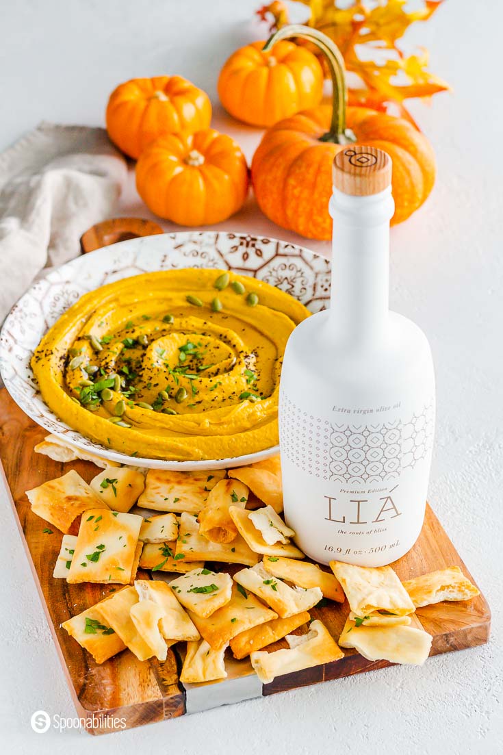 A white bottle with LIA extra virgin olive oil on top of a wooden board. Next to LIA EVOO some pita chips and in the background a large plate with pumpkin hummus and small pumpkins as a décor. Shop LIA at Spoonabilities.com