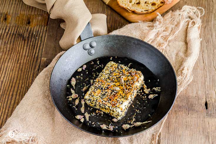 Close up photo of a pan-fried feta cheese with sesame crust garnished with thyme flowers. The pan is on top of a burlap napkin. Recipe at Spoonabilities.com
