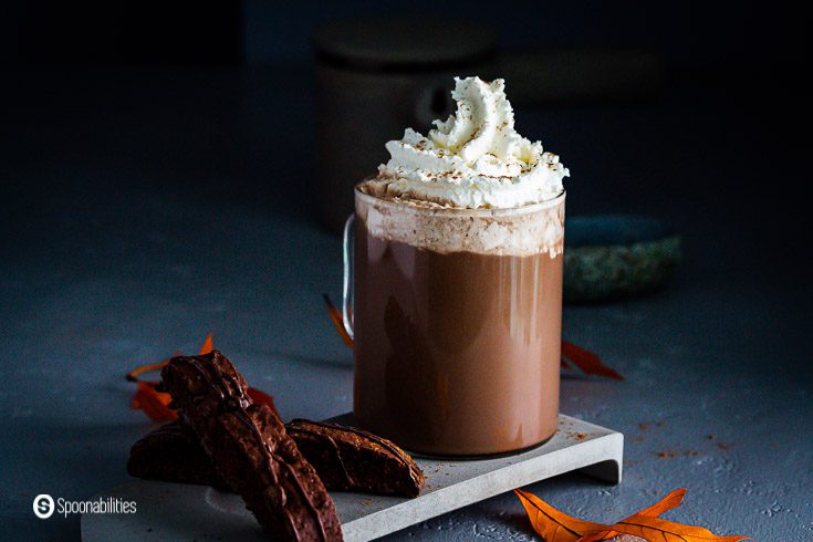One glass cup with pumpkin spice mocha topped with whipped cream. Next to the cup two chocolate biscotti and fall leaves around the glass cup. Recipe at Spoonabilities.com