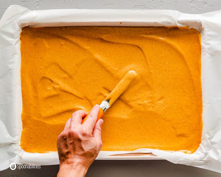 Pumpkin cake batter in a lined baking tray and a hand flattering out the batter using an offset spatula. Recipe at Spoonabilities.com
