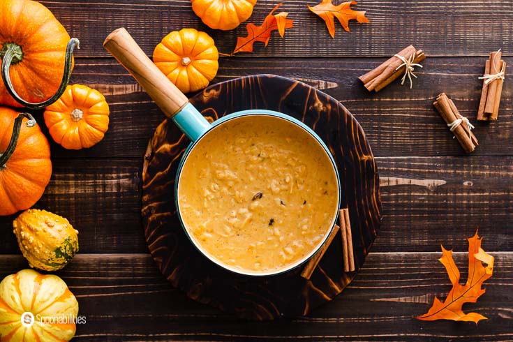 An overhead photo with a teal color pot with a wooden handle. The pot has pumpkin rice pudding with coconut milk and maple syrup. The table is decorated with mini pumpkins and fall leaves. Recipe at Spoonabilities.com