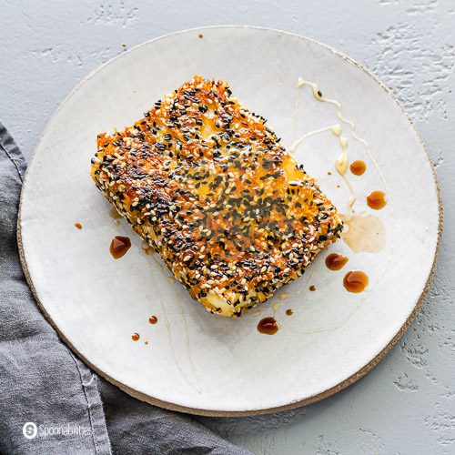 A round white plate with a block of sesame seeds crusted pan seared feta with a drizzle of honey and balsamic vinegar. Recipe at Spoonabilities.com