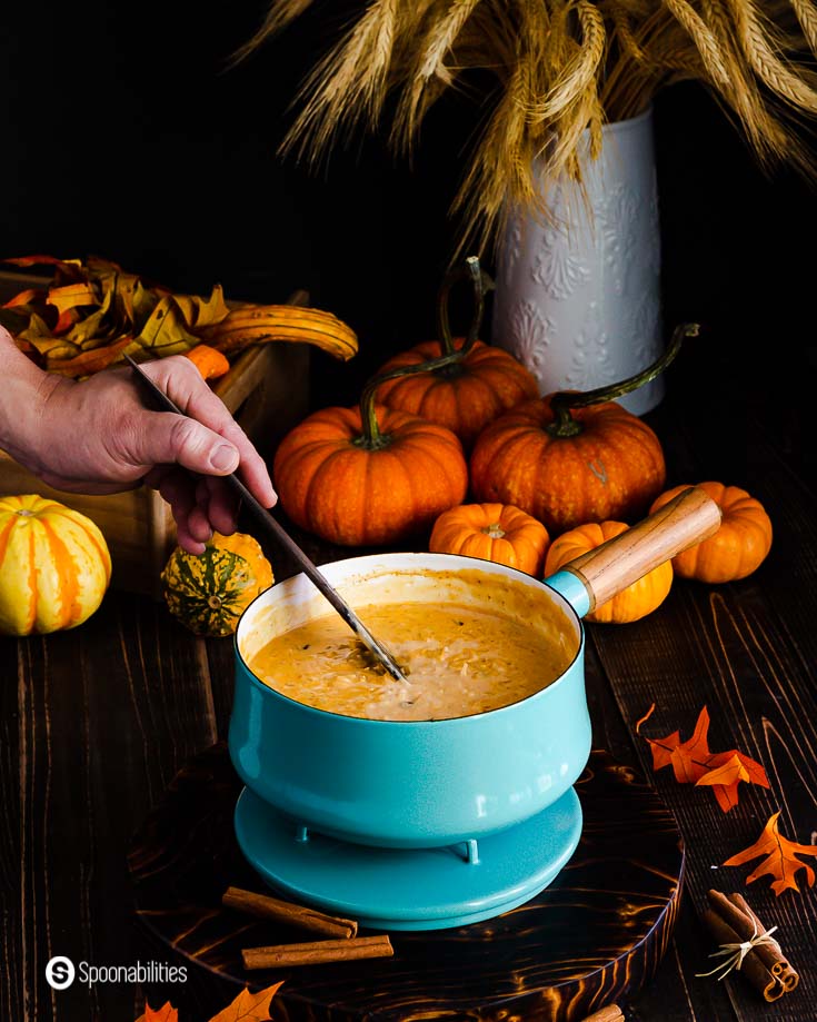 A pot with a wooden handle in a teal color with pumpkin rice pudding with coconut milk and Vermont maple syrup. One hand stirring the pot. Recipe at Spoonabilities.com