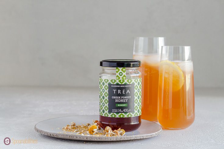 A jar of Greek honey on top of a round white plate and two glasses in the background with punch. The Trea Greek Forest Honey is available at Spoonabilities.com