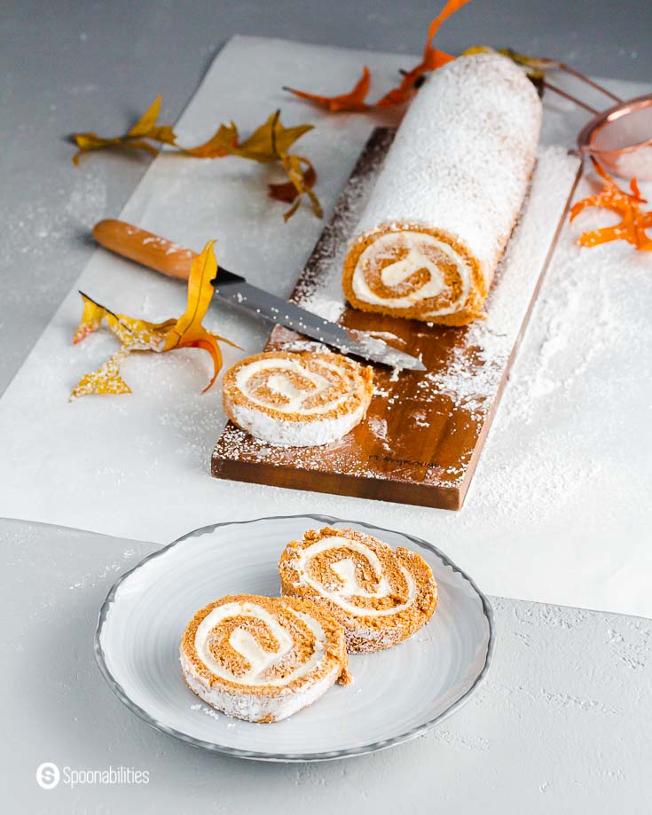 Two slices of pumpkin cake on a grey plate. In the background, the rest of the pumpkin roll on a wooden board with a piece on the board. Recipe at Spoonabilities.com