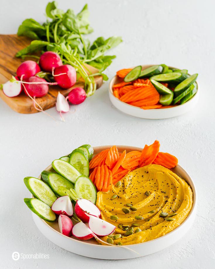 A large plate with some hummus with pumpkin flavor. Serve it with fresh cucumbers, radishes, and carrots. In the back ground two more plates with vegetables. Recipe at spoonabilities.com