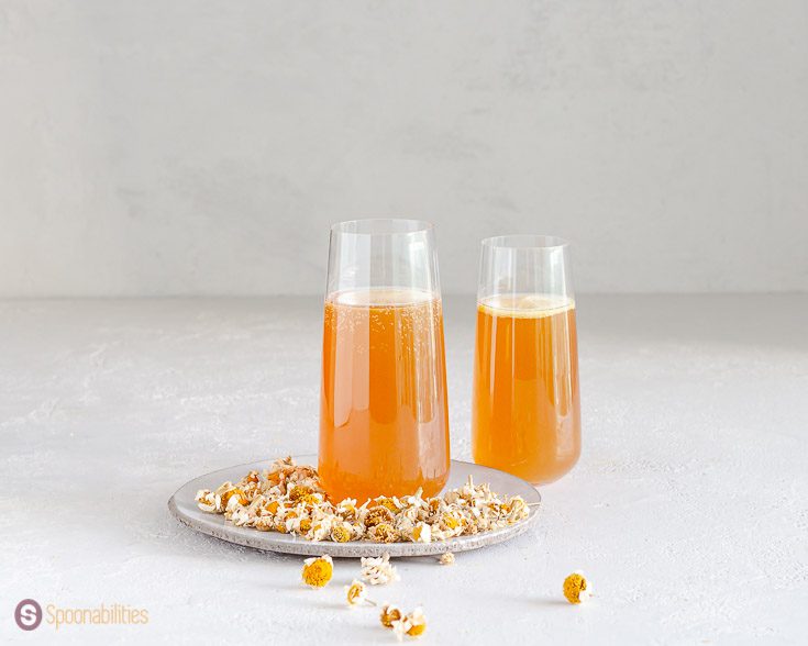 A flute glass on top of a plate with chamomile flowers around the plate. A second glass is on the right side. The glasses have a cocktail made out of pear infused vodka, chamomile tea, and honey syrup. Recipe at Spoonabilities.com