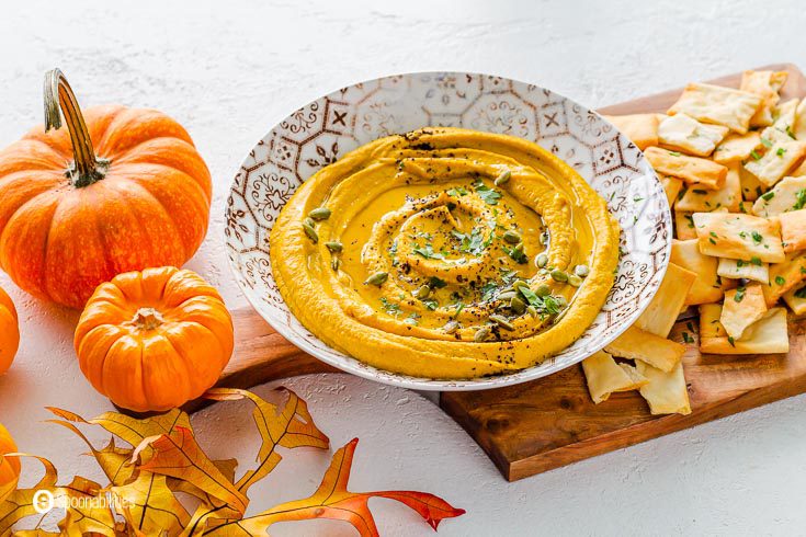 Large bowl with pumpkin hummus garnished with pumpkin seeds and chopped parsley. The plate is on top of a wooden board with pita chips on the right side and on the left side of the plate some mini pumpkins and fall leaves. Recipe at Spoonabilities.com
